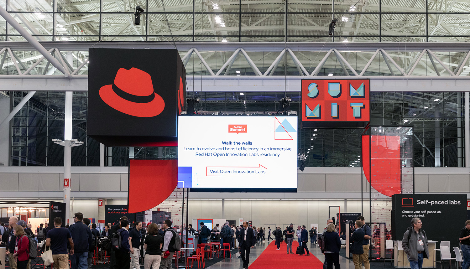 Foto scattata nella sala espositiva del Red Hat Summit. Un cartellone di grandi dimensioni con l'immagine del cappello è appeso sopra gli stand.