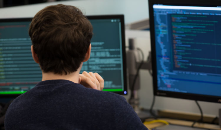 Eine technische Fachkraft arbeitet an einem Schreibtisch mit Computermonitoren, auf denen Code angezeigt wird